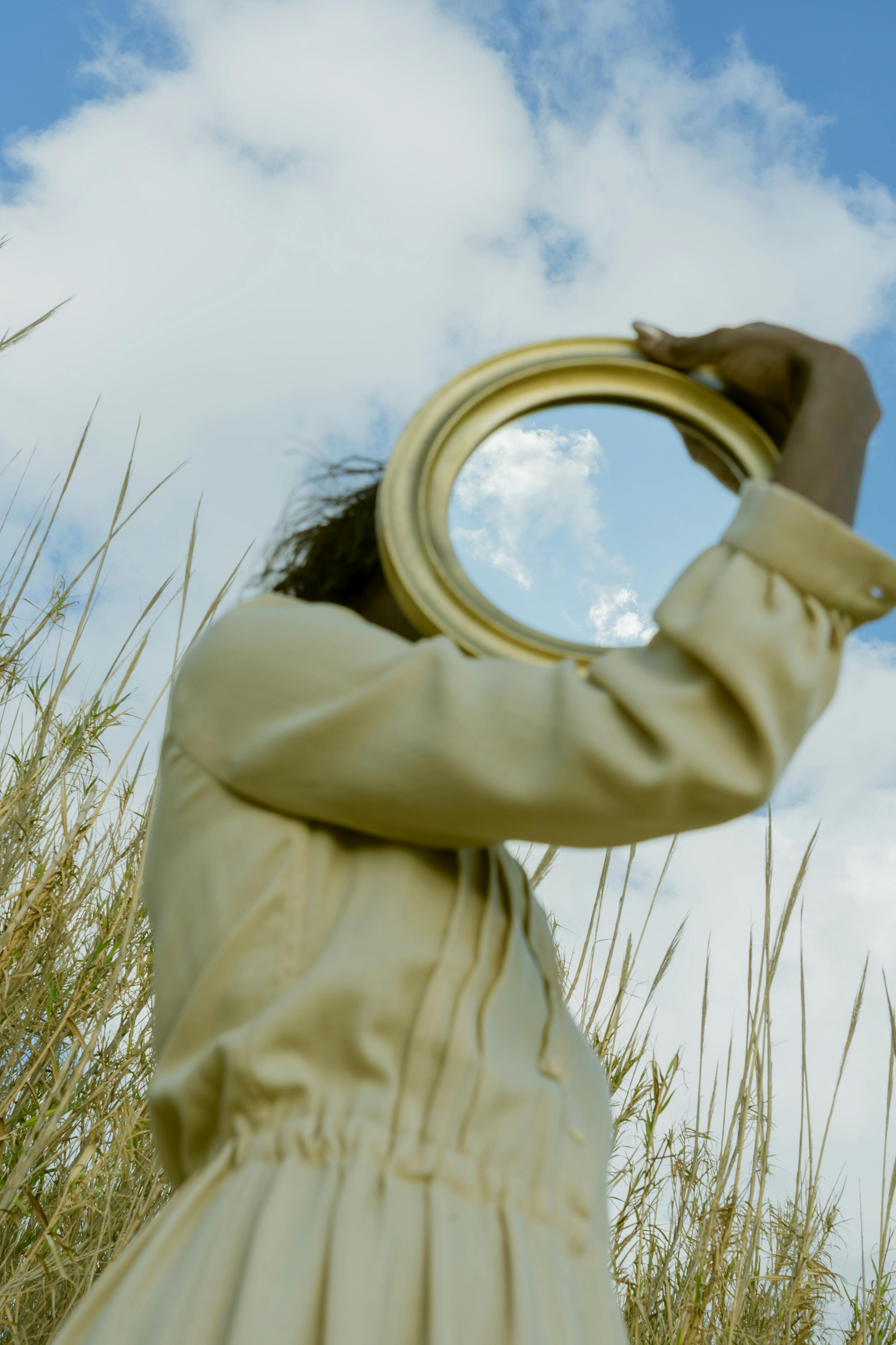 Person holding a round mirror that reflects the sky, obscuring their face in a tall field of grass